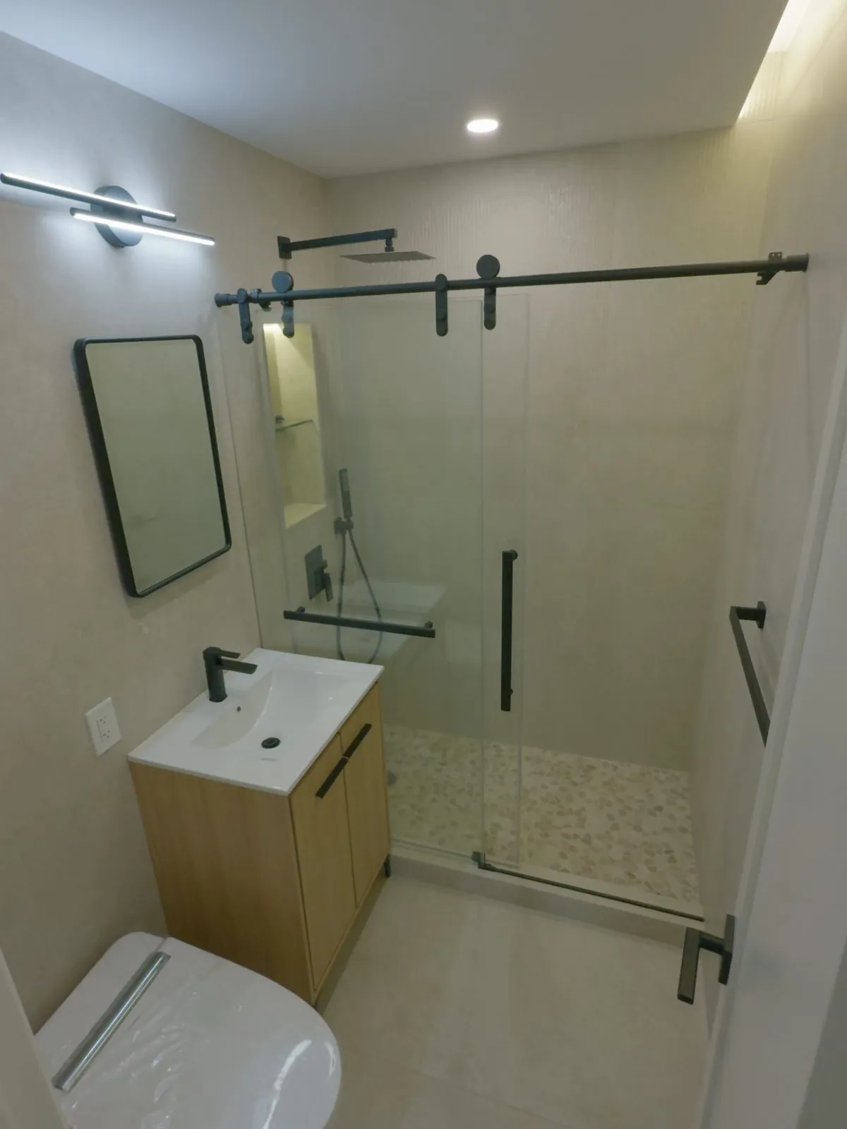 Finished modern bathroom with a wood vanity, glass shower and pebble floor