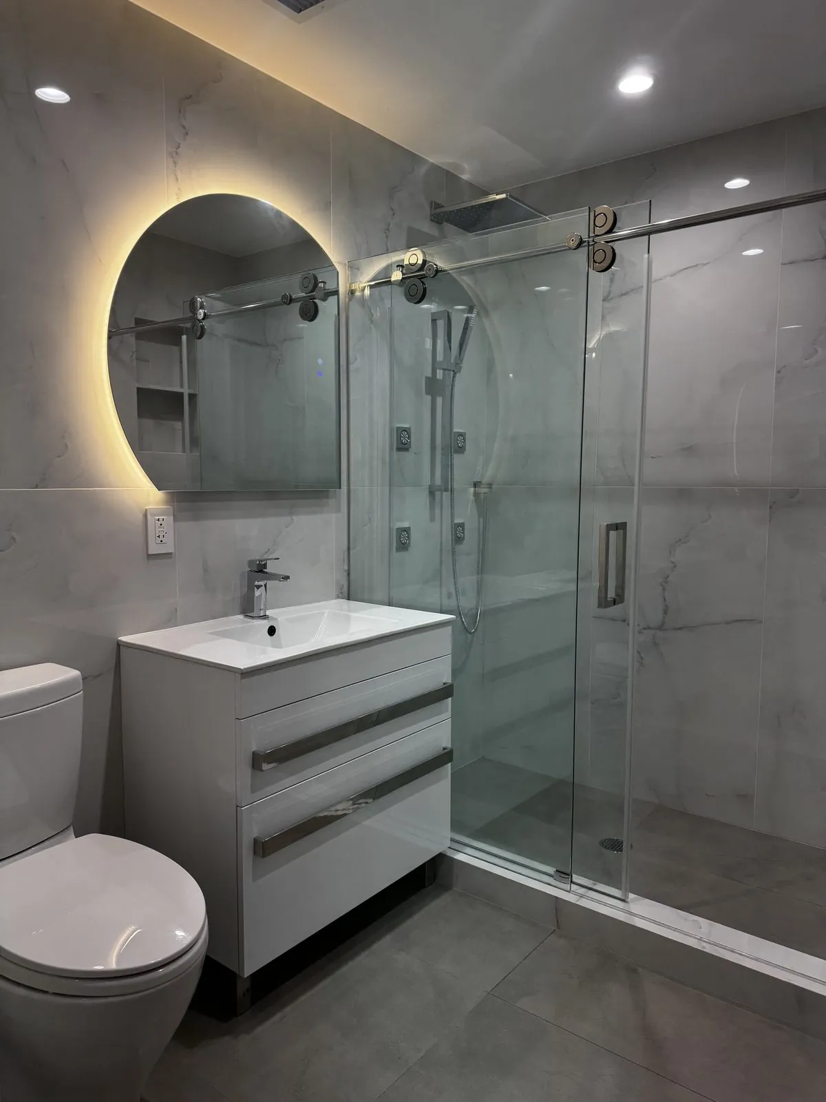 Finished gray marble bathroom with a backlit round mirror, floating vanity and frameless glass shower