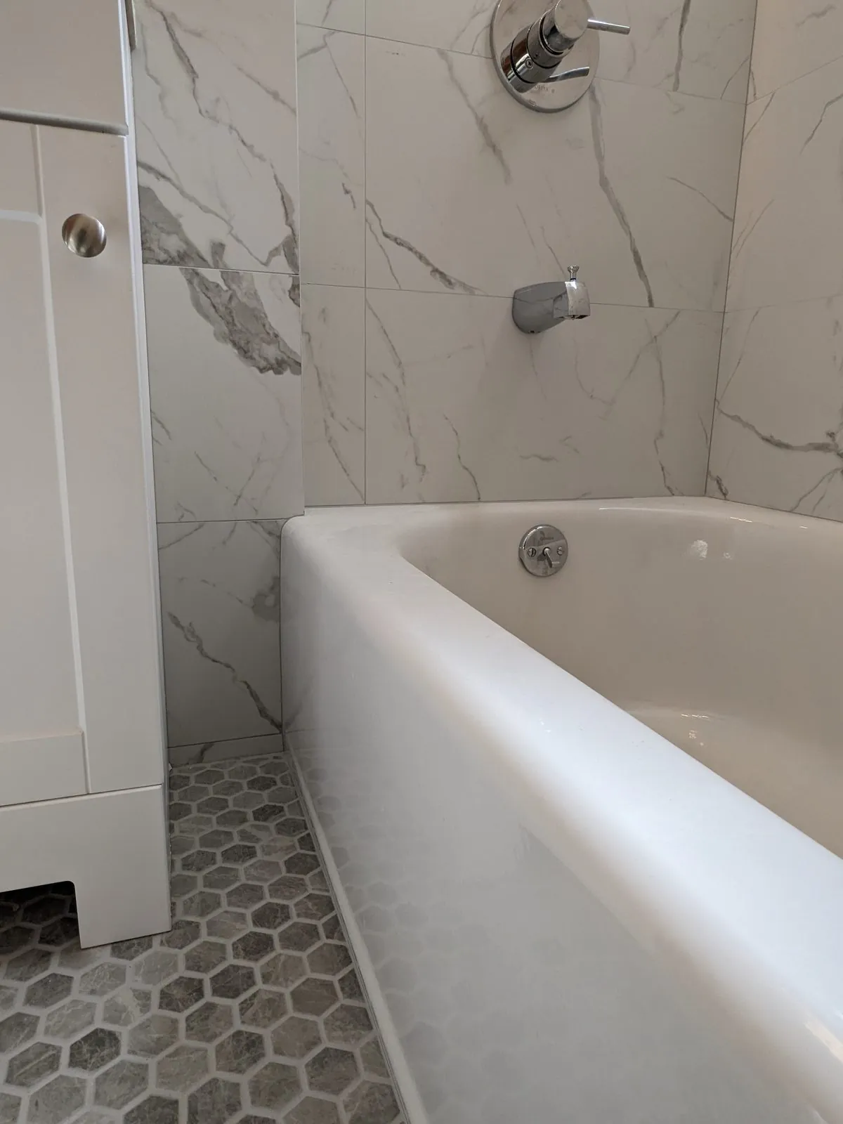 Rebuilt bathroom with white marble tub surround, chrome valve and gray marble hex floor