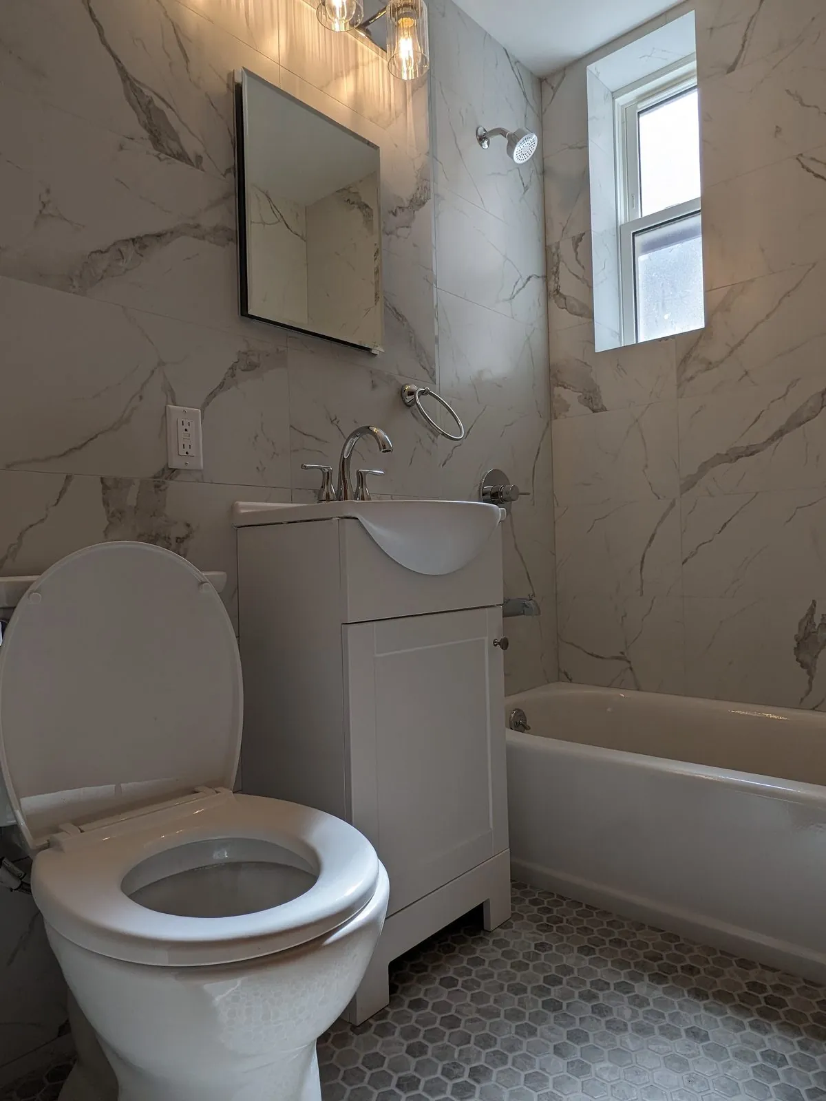 Finished marble bathroom: vanity with bulb light bar and mirror, tub under the window, gray hex floor